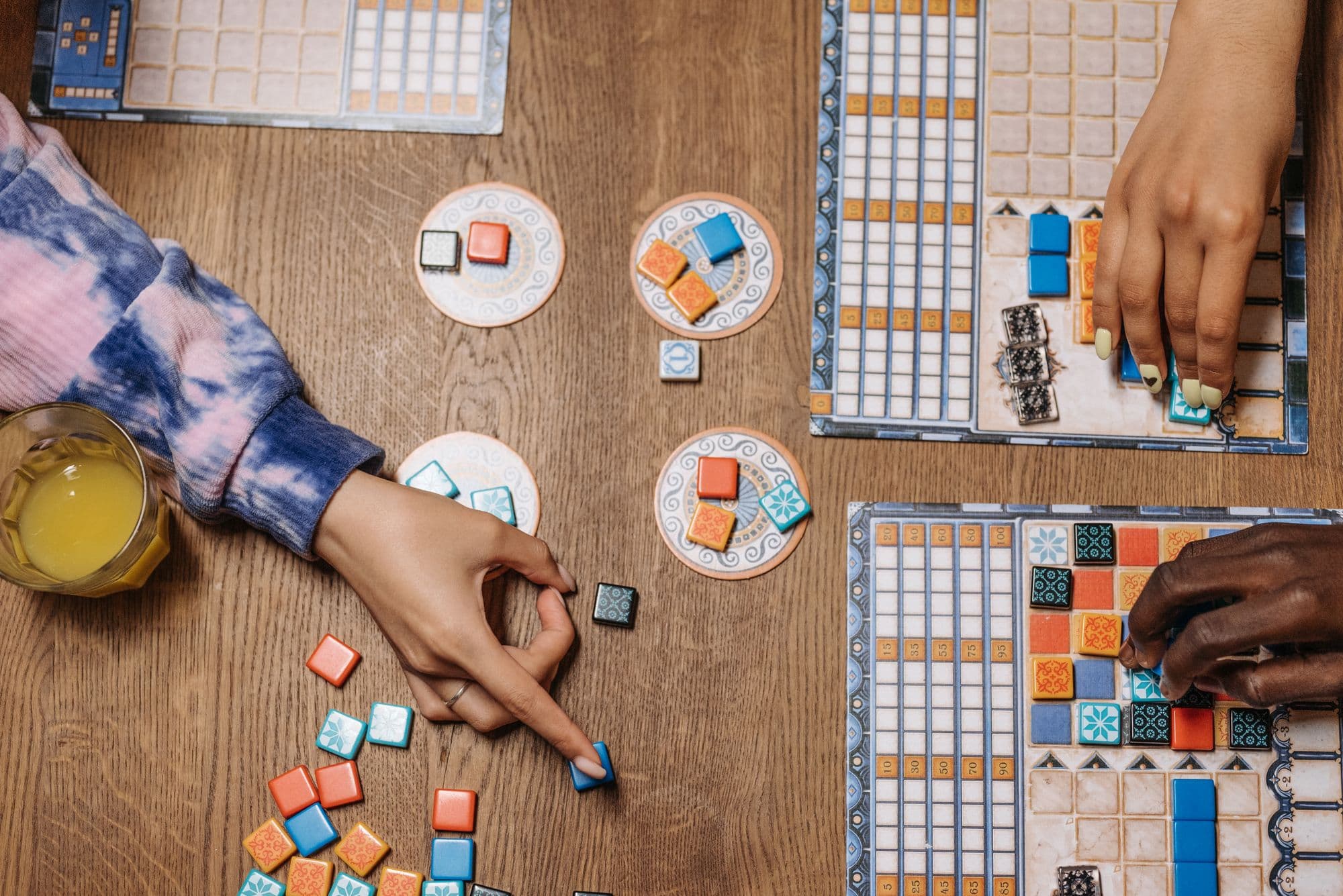 Overhead view of two people mid-game at a tile-laying board game, a drink beside the boards